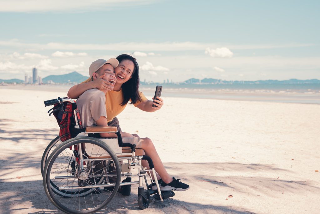 Happy mother and disabled teenage boy on wheelchair use social media, Sea beach background.